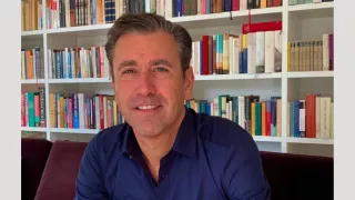 A man in a blue shirt sitting in front of a bookcase