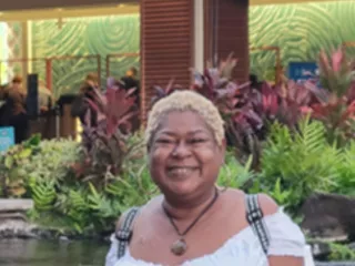 A woman stands in front of a fountain with plants, smiling at the camera. Tropical plants are visible in the background.