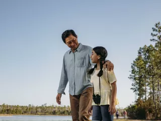 A father and daughter walking by a lake, the daughter wearing binoculars around her neck