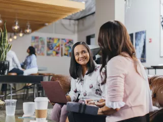 Two women sitting on a coffee shop sofa, engaged in conversation, while one woman uses a laptop